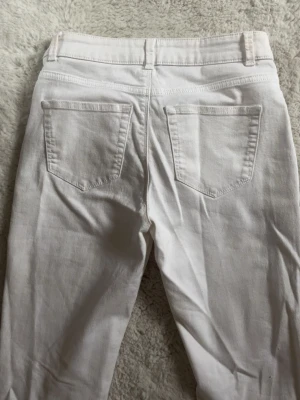 Vita bootcut jeans - Säljer ett par vita bootcut jeans med klassisk femficksdesign och bälteshällor. Jeansen har en snygg utsvängd passform och är tillverkade i ett mjukt bomullsmaterial med lite stretch för extra komfort.