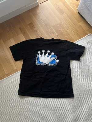 Svart Stüssy t-shirt med krona - Svart t-shirt från Stüssy med stor grafisk krona i vitt och blått på ryggen och mindre krona med Stüssy-logga på bröstet. Klassisk passform och rund hals. Tillverkad i mjuk bomull, perfekt för en streetwear-look. DU VET VARFÖR DEN ÄR BILLIG!