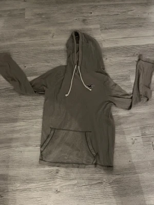 Grå hoodie från Hollister med logga - Snygg grå hoodie från Hollister med vit dragsko i huvan och klassisk Hollister-logga på bröstet. Hoodien har en stor magficka och är tillverkad i ett mjukt, ribbat material som känns skönt mot huden. Perfekt för en avslappnad och trendig look.