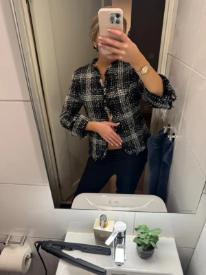 Svartvit tweedblus med fransar - Trendig svartvit blus i tweed med rutigt mönster och råa fransade kanter. Blusen har lång ärm och en klassisk krage, perfekt för en cool och stilren look. Passar till både jeans och kostymbyxor.