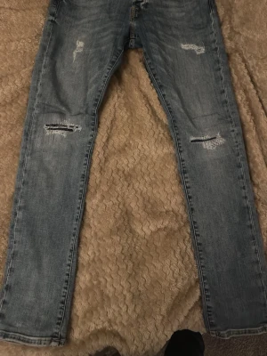 Jack and Jones  - Säljer ett par blå  jeans med slitningar och hål på backen men det är väldigt litet och knappt syns man kan fixa det enkelt . Jeansen har klassisk femficksdesign och är i ett mjukt denimtyg som ger en avslappnad vibe. Perfekta för dig som gillar en trendig och casual stil.