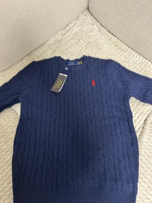Mörkblå kabelstickad tröja Polo Ralph Lauren - Mörkblå kabelstickad tröja från Polo Ralph Lauren med rund halsringning och röd broderad logga på bröstet. Tröjan har ribbade muddar vid ärmslut och nederkant. Perfekt för en stilren och klassisk look.