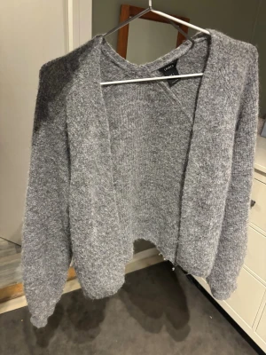 Grå stickad cardigan från LINDEX - Mysig grå stickad cardigan från LINDEX med öppen front och avslappnad passform. Tröjan har långa ärmar och är gjord i ett fluffigt, mjukt material som känns skönt mot huden. Perfekt att slänga över en t-shirt eller topp för extra värme.