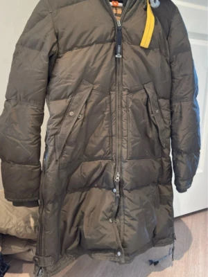 Mörkgrön lång dunjacka Parajumpers - Lång, mörkgrön dunjacka från Parajumpers med quiltad design och ikonisk gul rem vid halsen. Jackan har dragkedja framtill, stora fickor med knappar och en varm, vadderad känsla. Perfekt för kalla vinterdagar.