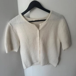 Vit kortärmad kofta från Gina Tricot - Söt vit kofta från Gina Tricot med korta ärmar och rund halsringning. Koftan har en mjuk, fluffig känsla och knäpps med små vita knappar framtill. Perfekt att slänga över en topp för en trendig look.