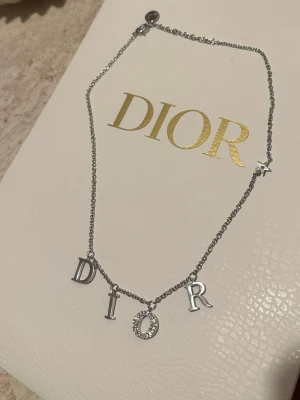  Dior med bokstäver silver  - Snyggt halsband från Dior i silverfärgad metall med hängande bokstäver som stavar DIOR. O:et är dekorerat med små gnistrande stenar och på kedjan finns även en liten stjärnberlock. Perfekt för dig som gillar exklusiva accessoarer med statement-känsla.