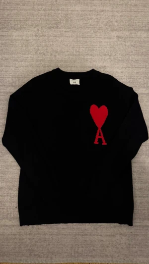 Ami Paris Tröja - Ami Paris svarta stickade tröja med ikoniskt rött hjärta och bokstaven A broderat på bröstet. Rund halsringning och långa ärmar ger en stilren look med en modern twist.