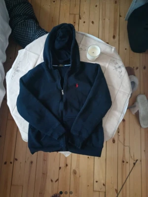 Marinblå hoodie från Polo Ralph Lauren - Snygg marinblå hoodie från Polo Ralph Lauren med dragkedja framtill och klassisk röd logga på bröstet. Mjuk insida och huva för extra komfort. Perfekt för en avslappnad och stilren look. Tillverkad i bomullsmix.