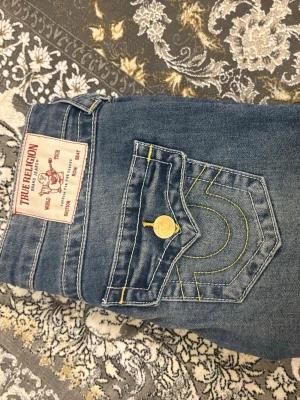 True Religion Joey Low Rise Flare Jeans - Säljer ett par helt nya blå True Religion Joey jeans med låg midja och flare-ben. Ingen täcken på användning för de är helt nya, fick i present men för småa. Klassisk gul kontrastsöm, ficklock med knapp och ikonisk logga på bakfickan. Tillverkade i slitstarkt jeansmaterial. Perfekta för dig som gillar statement-jeans med unik design. PRIS KAN DISKUTERAS 