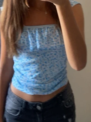 Blommig blå linnetopp - Söt croppad linnetopp i ljusblått med små blommor och volangdetalj framtill. Toppen har ärmlös design och fyrkantig ringning, perfekt för varma dagar. Materialet känns mjukt och lätt mot huden.