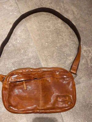 Brun axelväska i skinn från Gusti Leder - Snygg brun axelväska från Gusti Leder i mjukt skinn. Väskan har ett justerbart axelband i tyg, dragkedja upptill och flera fack både fram och bak. Perfekt storlek för det viktigaste och en stilren look till alla outfits.