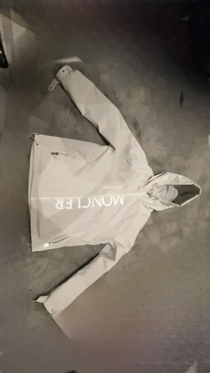 Grå vindjacka med huva från Moncler - Snygg grå vindjacka från Moncler med stor logga framtill och klassisk patch på ärmen. Jackan har huva med dragsko, dragkedja framtill och två sidofickor med dragkedja. Perfekt för blåsiga dagar och enkel att matcha till olika outfits.