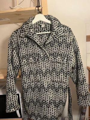 Svartvit mönstrad kappa med bälte - Snygg svartvit kappa med grafiskt mönster och bälte i midjan. Kappan har bred krage, långa ärmar och två bröstfickor. Perfekt för dig som vill sticka ut med en trendig och unik look.