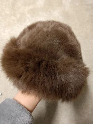 Brun pälsmössa i klassisk stil - Säljer en fluffig och varm brun pälsmössa med rund form. Mössan har en lyxig känsla och är perfekt för kalla dagar. Materialet är mjukt och tätt, vilket ger en snygg och trendig look. Passar dig som vill sticka ut med en klassisk accessoar.