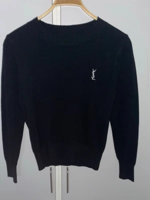 Svart stickad tröja från Yves Saint Laurent - Klassisk svart stickad tröja från Yves Saint Laurent med diskret vitt YSL-broderi på bröstet. Storlek S, tröjan har rund halsringning, långa ärmar och ribbade muddar vid ärmslut och nederkant. Perfekt för en stilren och tidlös look.