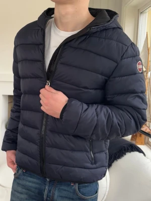 Mörkblå pufferjacka från Colmar - Snygg mörkblå pufferjacka från Colmar med huva och dragkedja framtill. Jackan har quiltad design, två fickor med dragkedja och en logotyp på ärmen. Perfekt för kyliga dagar och enkel att matcha med jeans eller hoodies.