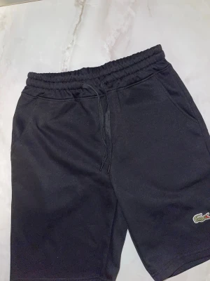Svarta shorts från Lacoste med snörning - Svarta shorts från Lacoste med elastisk midja och snörning framtill. Klassisk krokodillogo på vänster ben. Tillverkade i mjukt material, perfekta för chill dagar eller träning. Två sidofickor för det viktigaste.