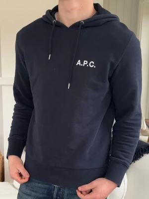 Mörkblå hoodie från A.P.C. - Snygg mörkblå hoodie från A.P.C. med vit logga på bröstet. Klassisk modell med huva och dragsnören, ribbade muddar vid ärmslut och nederkant. Tillverkad i mjukt bomullsmaterial som känns skönt mot huden. Perfekt för en avslappnad och stilren look.