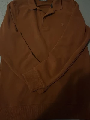 Brun långärmad tröja Polo Ralph Lauren - Snygg brun långärmad tröja från Polo Ralph Lauren med klassisk pikékrage och diskret broderad logga på bröstet. Tröjan har ribbade muddar vid ärmslut och nederkant, perfekt för en stilren och avslappnad look.