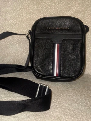 Svart axelväska Tommy Hilfiger - Snygg svart axelväska från Tommy Hilfiger i strukturerat konstläder. Väskan har justerbar axelrem och en ikonisk rand i vitt, rött och blått på framsidan. Perfekt storlek för det viktigaste och stilren design med diskret logga upptill.