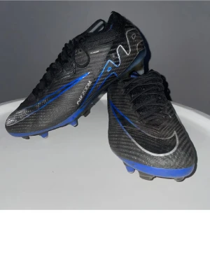 Nike Mercurial svarta fotbollsskor - Snygga svarta Nike Mercurial fotbollsskor med blå och silverdetaljer. Skorna har snörning och är tillverkade i syntetmaterial för bästa grepp och känsla på planen. Perfekta för dig som vill ha stilrena och lätta skor till fotbollsträningen.