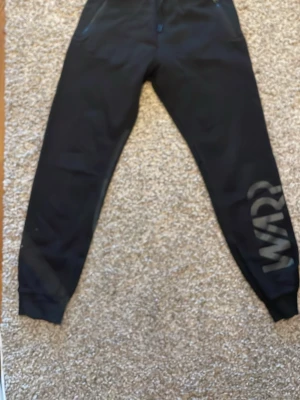 Svarta mjukisbyxor med dragkedjor - Säljer ett par svarta mjukisbyxor med snörning i midjan och dragkedjor på fickorna. Ena benet har en stor tryckt text med 'WRP' i mörk ton. Perfekta för chill eller träning, riktigt sköna och stilrena.