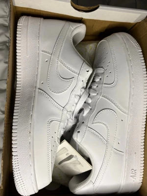 Nike Air Force 1 vita sneakers - Klassiska helvita Nike Air Force 1 sneakers med låg profil. Skorna har en rund tå, snörning och är tillverkade i slätt skinn med diskreta sömmar och Nike-logga på sidan och hälen. Perfekta för dig som gillar stilrena och tidlösa sneakers.