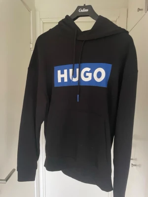 Svart hoodie från Hugo med blå logga - Svart hoodie från Hugo med stor blå logga på bröstet och vit text. Hoodien är använd ett fåtal gånger. Storlek S