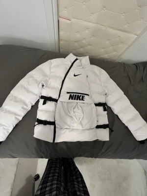Vit pufferjacka från Nike - Säljer en riktigt snygg vit pufferjacka från Nike med svarta detaljer och stor logga på bröstet. Jackan har justerbara spännen i midjan, svart innerfoder och dragkedja i sned design. Perfekt för dig som vill sticka ut med sportig stil.