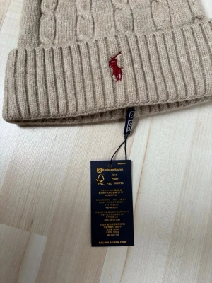 Beige kabelstickad mössa Polo Ralph Lauren - Snygg beige kabelstickad mössa från Polo Ralph Lauren med uppvikt kant och klassisk röd broderad logga framtill. Tillverkad i mjuk ullblandning som håller dig varm och stilren under kalla dagar. Perfekt accessoar för en trendig look.