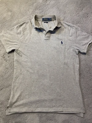 Grå piké från Polo Ralph Lauren - Klassisk grå pikétröja från Polo Ralph Lauren med korta ärmar och blå logga broderad på bröstet. Tröjan har krage och knappar vid halsen. Perfekt till en avslappnad men ändå stilren look. Mitt pris 280kr. Säljer pga för stor.