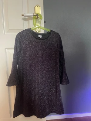 Glittrig svart klänning Lindex Kids - Supersnygg svart glittrig klänning från Lindex Kids med trekvartsärm och volang vid ärmslut. Perfekt för dig som vill sticka ut lite extra med en enkel men ändå festlig look. Mjuk och stretchig syntetmix som känns skön att bära.