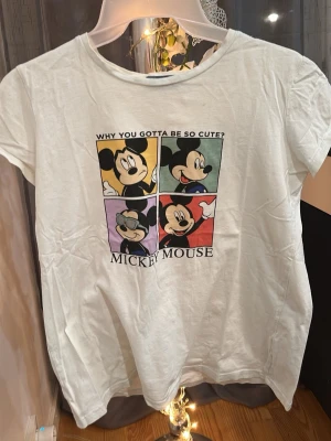 Vit Mickey Mouse t-shirt från Disney - Vit t-shirt från Disney med färgglatt tryck av Mickey Mouse i fyra rutor och texten 'WHY YOU GOTTA BE SO CUTE?'. Rund halsringning och korta ärmar. Perfekt för dig som gillar klassiska Disney-figurer och vill ha en lekfull stil.
