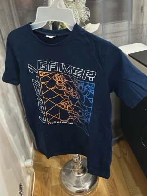 Blå gamer t-shirt från LC Waikiki - Cool blå t-shirt från LC Waikiki med gamer-tryck och texten 'LEVEL 1 GAMER' på framsidan. Motiv med spelkontroller i orange och blått. Perfekt för dig som älskar gaming och vill sticka ut med en snygg och bekväm t-shirt.