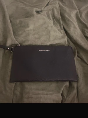 Svart kuvertväska från Michael Kors - Stilren svart kuvertväska från Michael Kors i slätt skinn med silverfärgad logga framtill. Väskan har en avtagbar handledsrem och dragkedja upptill. Perfekt för att bära det viktigaste och ger en clean look till din outfit.