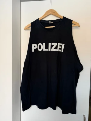 Svart ärmlös topp med POLIZEI-tryck - Cool svart ärmlös topp med stort vitt POLIZEI-tryck fram och mindre tryck bak i nacken. Oversized passform och breda ärmhål ger en avslappnad look. Perfekt för dig som vill sticka ut med en statement-topp.