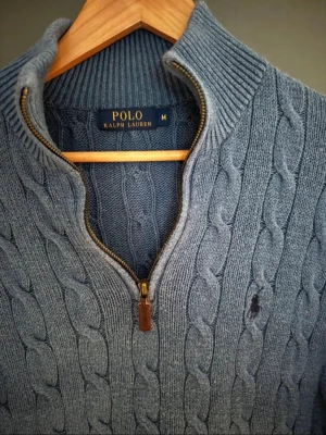 Blå kabelstickad tröja Polo Ralph Lauren - Kabelstickad blå tröja från Polo Ralph Lauren med halv dragkedja och hög krage. Tröjan har ribbstickade muddar och nederkant samt den klassiska broderade loggan på bröstet. Perfekt för lager-på-lager och snygg till jeans.