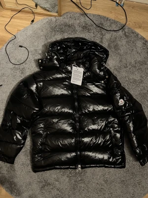 Svart glansig dunjacka från Moncler - Snygg svart dunjacka från Moncler med glansig finish och klassisk pufferdesign. Jackan har huva, dragkedja framtill och Moncler-logga på ärmen. Perfekt för kalla dagar och ger en riktigt modern look. NFC tagg fungerar 