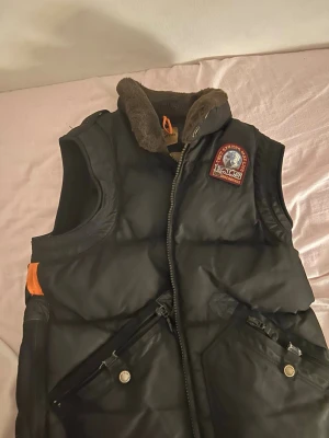 Svart dunväst från Parajumpers - Svart dunväst från Parajumpers med brunt foder i kragen och orange detaljer. Västen har två snedställda fickor med dragkedja och tryckknappar samt ett stort broderat märke på bröstet. Perfekt för lager-på-lager och streetstyle.