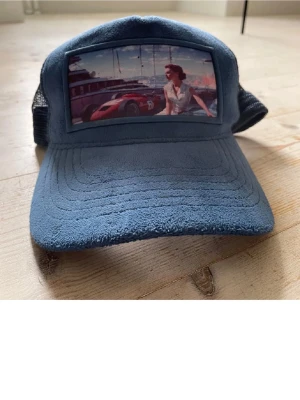 Blå meshkeps med retrotryck - Säljer en blå keps med mesh på sidorna och baksidan. Framtill finns ett retroinspirerat tryck med bil och kvinna. Kepsen har böjd skärm och är tillverkad i mjukt tyg framtill och mesh bak för extra ventilation. Perfekt för dig som gillar unik streetwear.