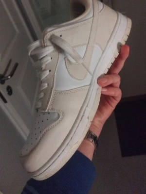 Nike Dunk Low vita sneakers - Nike Dunk Low sneakers i vitt och ljusbeige skinn med klassisk rund tå och snörning. Swoosh-logga på sidan och platt sula. Perfekta för dig som gillar stilrena och tidlösa sneakers.