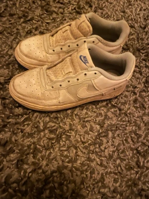 Nike Air Force 1 vita sneakers - Klassiska Nike Air Force 1 sneakers i vitt skinn med blå detaljer och blå sula. Modellen har rund tå, snörning och den ikoniska swoosh-loggan på sidan. Perfekta för dig som gillar streetstyle och tidlös design.