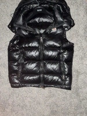 Svart dunväst från moncler med huva - Snygg svart dunväst från moncler med glansig finish och avtagbar huva. Västen har två dragkedjeförsedda fickor framtill och en diskret logga på bröstet. Perfekt för lager på lager och ger en cool streetwear-look.