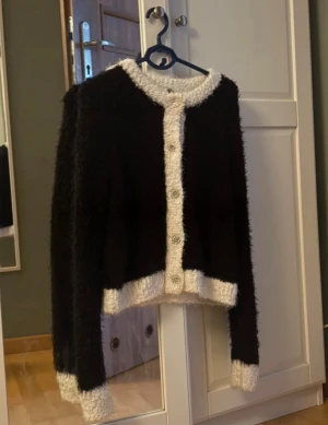 Svart fluffig kofta med pärlknappar - Fluffig svart kofta med vita kanter och dekorativa pärlknappar. Koftan har lång ärm och är tillverkad i ett mjukt, stickat material som känns riktigt cozy. Perfekt för att lyfta din outfit med en chic touch.
