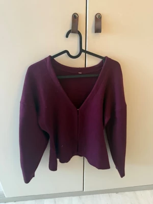 Vinröd kofta med v-ringning - Mysig vinröd kofta med djup v-ringning och lång ärm. Koftan har en enkel, stilren design utan synliga knappar eller fickor och är gjord i ett mjukt, tjockare material som passar perfekt till lager på lager. Perfekt för dig som gillar en avslappnad men snygg look. Ett litet hål men det syns inte men därav priset