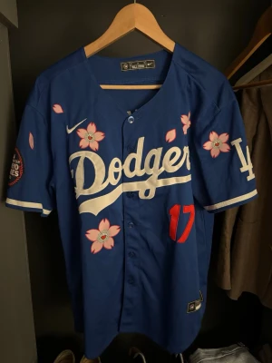 Blå Dodgers basebolltröja Nike M - Snygg blå Dodgers basebolltröja från Nike med vita och rosa körsbärsblommor, stort Dodgers-tryck på bröstet och röd siffra 17. Korta ärmar, knappar framtill och broderade detaljer. Perfekt för dig som gillar sportig stil och streetwear.