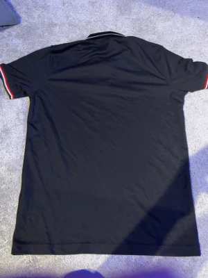 Svart Moncler pikétröja med ränder - Svart pikétröja från Moncler med ribbad krage och ärmslut. Ärmarna har röda och vita ränder som ger en sportig vibe. Tillverkad i mjuk bomull för skön känsla och klassisk passform.