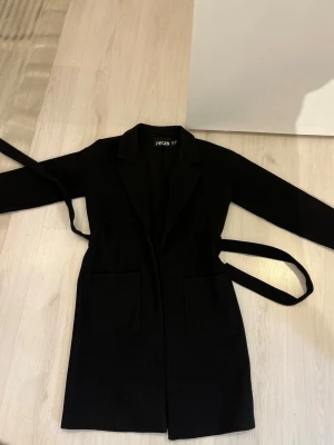 Svart kappa från Pieces med bälte - Stilren svart kappa från Pieces, köpt på boohoo. Två fickor framtill och ett matchande bälte i midjan. Klassisk krage och rak passform som passar till många olika looks. Perfekt för dig som gillar enkel och tidlös stil. Liten i storleken! 