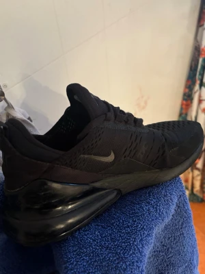 Svarta Nike Air Max 270 sneakers - Säljer ett par helsvarta Nike Air Max 270 sneakers med klassisk luftig mesh-överdel och stor synlig Air-enhet i hälen. Snygg och sportig design med diskret Nike-logga på sidan och snörning framtill. Perfekta för dig som gillar streetwear och sportig stil.