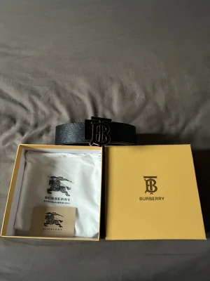 Svart Burberry bälte med monogramspänne - Snyggt svart bälte från Burberry med präglat monogrammönster längs hela remmen och ett stort metallspänne med Burberrys ikoniska TB-logga. Bältet kommer i originalförpackning med dustbag och certifikat. Perfekt accessoar för att lyfta din outfit.